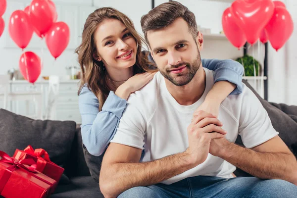 Happy young girlfriend embracing boyfriend in room with heart-shaped balloons — Stock Photo