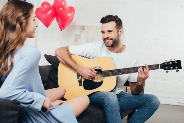 Joven tocando la guitarra a la novia sonriente con globos en forma de corazón en el fondo - foto de stock