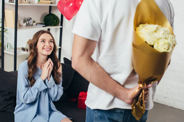 Young man holding bouquet of roses behind back while surprised smiling girl sitting with folded hands on sofa — Stock Photo