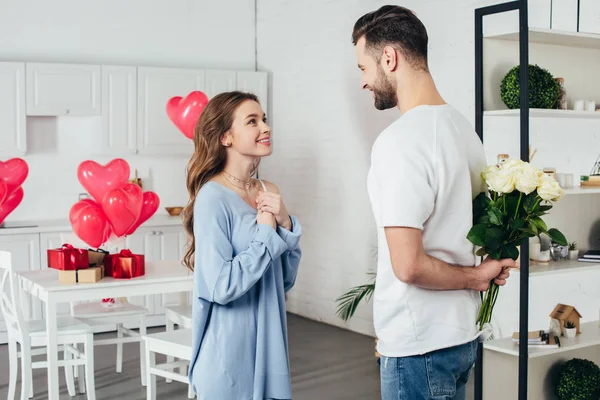 Young smiling girl in joyful expectation of st valentines day gift while smiling boyfriend holding roses bouquet behind back — Stock Photo