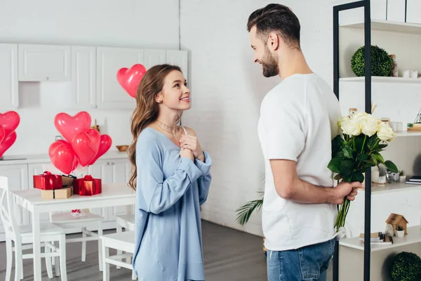 Jovem sorrindo menina na expectativa alegre de st dia dos namorados presente enquanto sorrindo namorado segurando rosas buquê atrás das costas — Fotografia de Stock