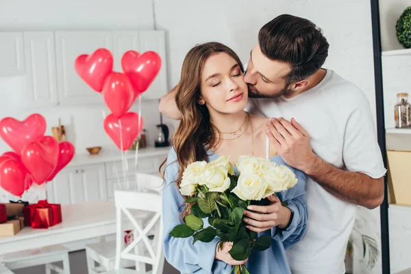 Happy girl holding roses bouquet with closed eyes while boyfriend kissing cheek and embracing girlfriend — Stock Photo