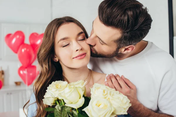 Selective focus of happy girl holding roses bouquet with closed eyes while boyfriend kissing cheek and embracing girlfriend — Stock Photo