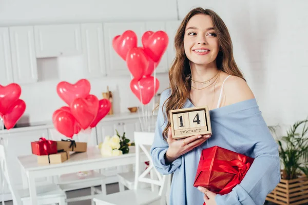 Happy smiling girl holding gift box and wooden calendar with st valentine day date at home — Stock Photo