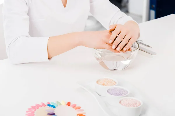 Cortado tiro de manicura en uniforme blanco sentado en el lugar de trabajo - foto de stock