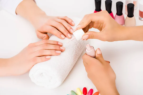 Vista cortada da menina com unhas curtas segurando as mãos na toalha enquanto manicure aplicando esmalte — Fotografia de Stock