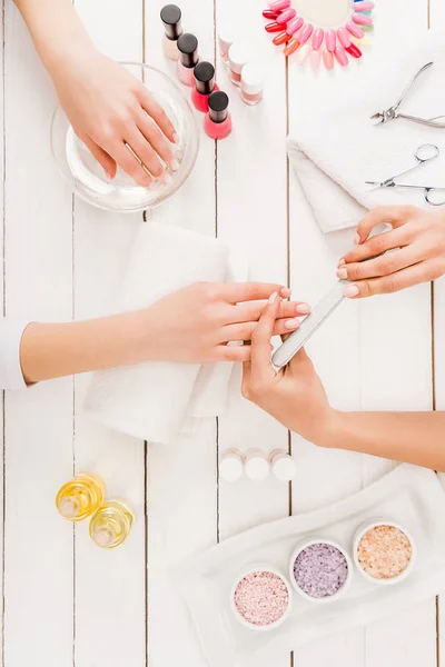 Visão superior da mulher mergulhando a mão na água enquanto manicure usando o arquivo de unhas — Fotografia de Stock