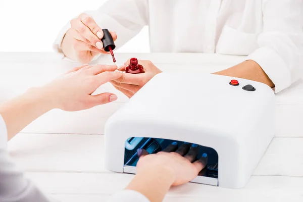 Cropped view of woman using nail lamp while manicurist painting fingernails — Stock Photo