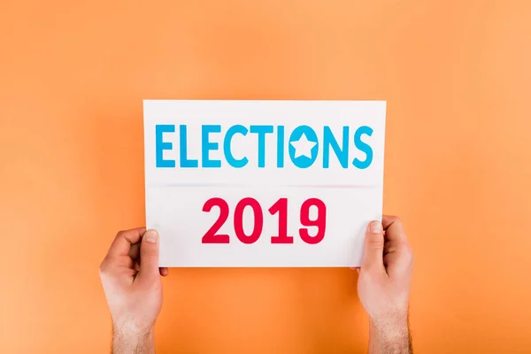Cropped view of man holding card with 'elections 2019' lettering isolated on orange — Stock Photo