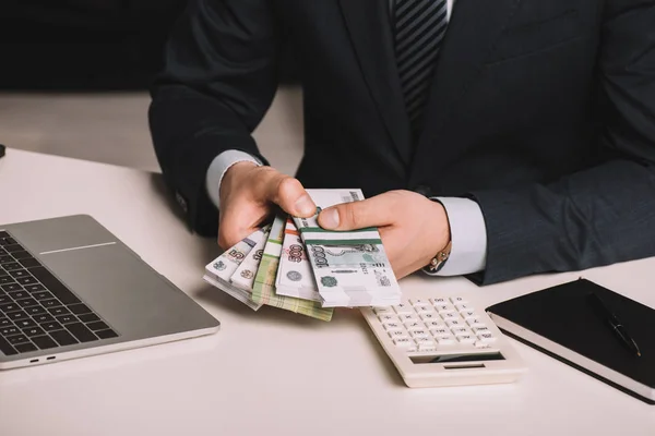 Обрезанный снимок бизнесмена с банкнотами российских рублей на рабочем месте с ноутбуком, калькулятором и ноутбуком — стоковое фото
