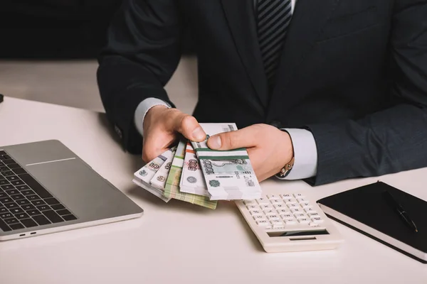 Colpo ritagliato di uomo d'affari in possesso di rubli russi banconote sul posto di lavoro con computer portatile, calcolatrice e notebook — Foto stock