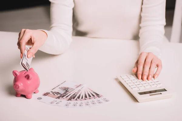 Cropped shot of woman using calculator and putting russian rubles into piggy bank — Stock Photo