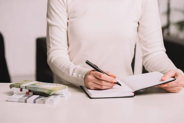 Recortado tiro de mujer escribiendo en cuaderno blanco sentado a la mesa con los billetes de rublos rusos - foto de stock