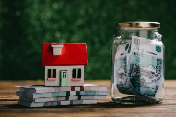 Vue rapprochée du bocal en verre avec de l'argent, modèle de maison et des billets de banque en roubles sur une table en bois — Photo de stock