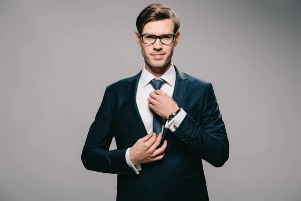 Красивый бизнесмен в костюме трогает галстук на сером фоне — стоковое фото