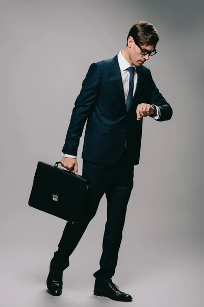 Serio uomo d'affari in possesso di valigetta e guardando orologio su sfondo grigio — Foto stock