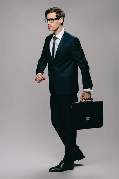 Uomo d'affari sicuro in giacca e cravatta camminare con valigetta su sfondo grigio — Foto stock