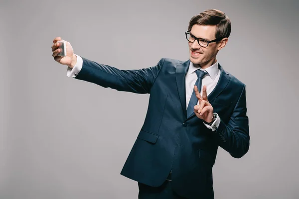 Cheerful businessman taking selfie and showing piece sign on grey background — Stock Photo