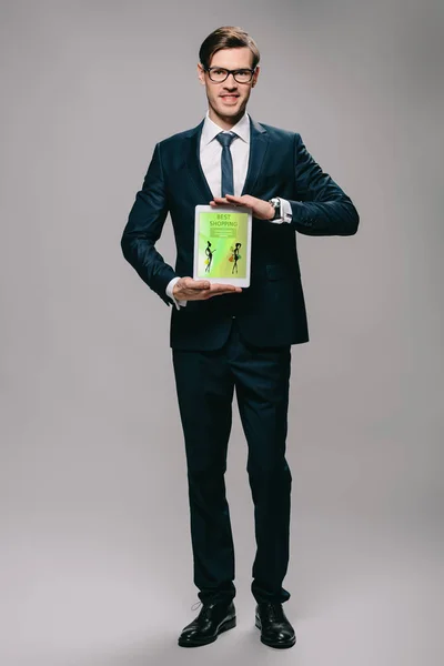 Красивий бізнесмен тримає цифровий планшет з найкращим додатком для покупок на екрані на сірому фоні — стокове фото