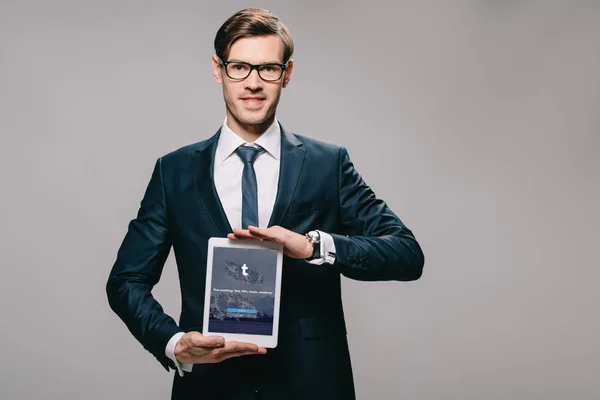 Cheerful businessman holding digital tablet with tumblr app on screen isolated on grey — Stock Photo