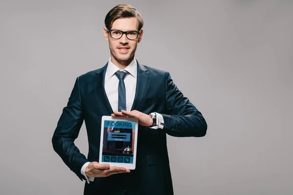 Счастливый бизнесмен, держащий цифровой планшет с приложением бронирования на экране изолированы на серый — стоковое фото
