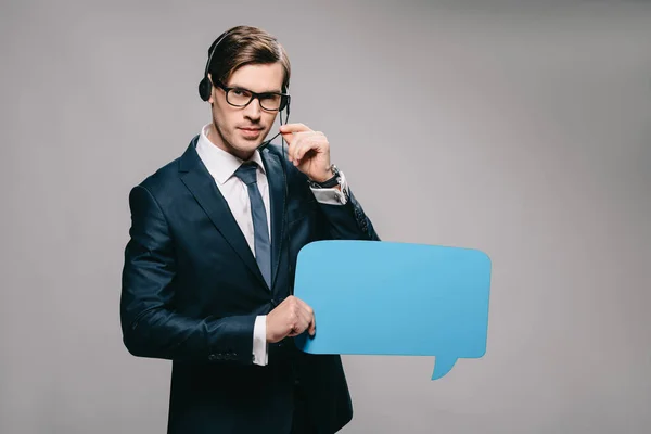 Serious man in suit wearing headset and holding speech bubble on grey background — Stock Photo