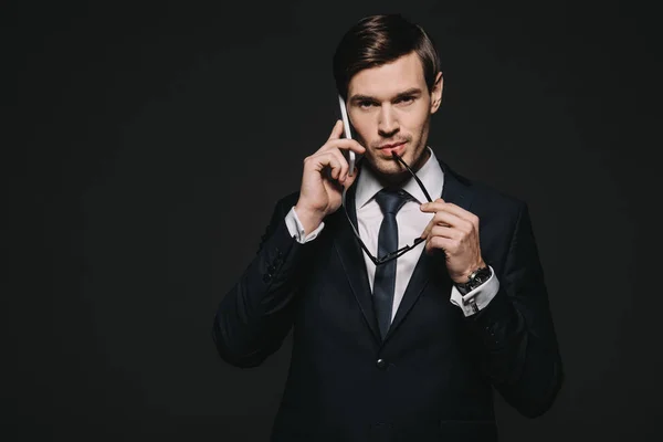 Handsome businessman talking on smartphone and holding glasses naer lips isolated on black — Stock Photo