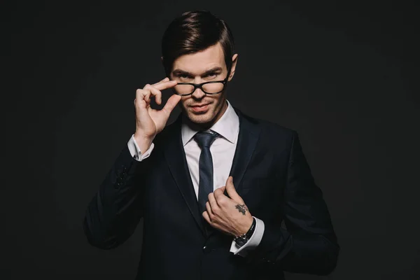 Guapo hombre de negocios tocando gafas mientras está de pie en traje aislado en negro - foto de stock
