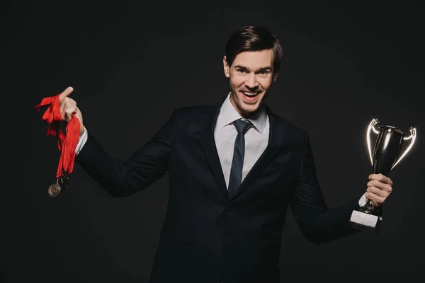 Happy businessman wearing medals and celebrating victory while holding trophy isolated on black — Stock Photo