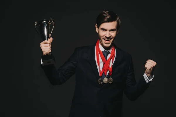 Homme d'affaires joyeux portant des médailles et célébrant la victoire tout en tenant trophée isolé sur noir — Photo de stock