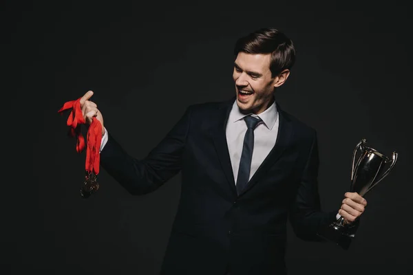 Cheerful businessman looking at medals and holding trophy isolated on black — Stock Photo
