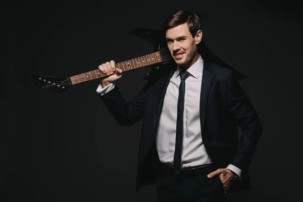 Guapo hombre de negocios sosteniendo la guitarra elecrtónica y de pie con la mano en el bolsillo aislado en negro - foto de stock