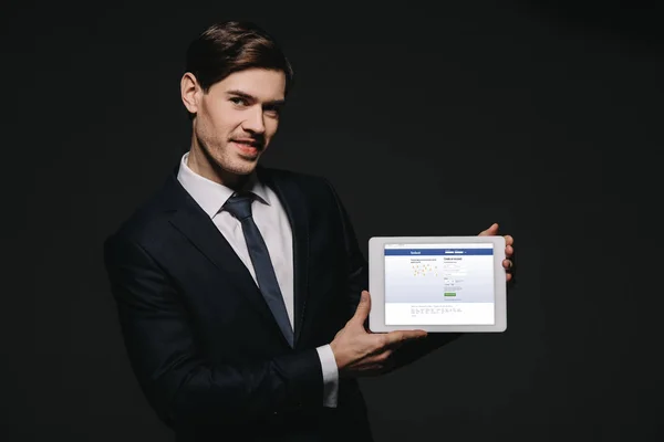 Cheerful businessman holding digital tablet with facebook app in screen isolated on black — Stock Photo