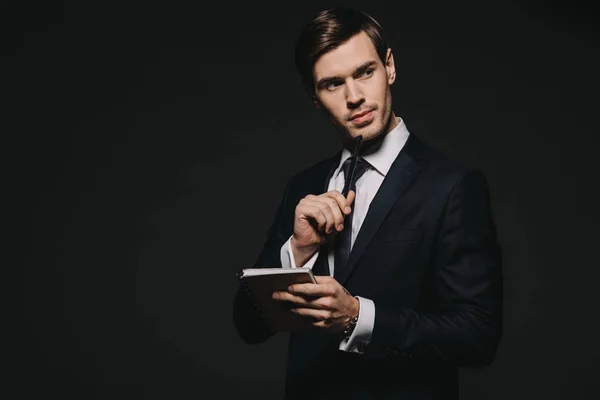 Pensativo hombre de negocios con cuaderno y pluma en las manos aisladas en negro - foto de stock
