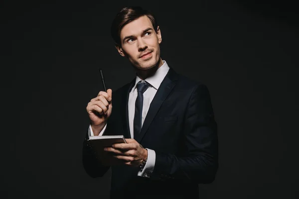 Persive businessman holding notebook and pen in hands isolated on black — Stock Photo