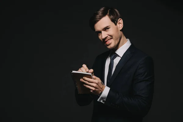 Empresário alegre escrevendo em notebook com caneta isolada em preto — Fotografia de Stock