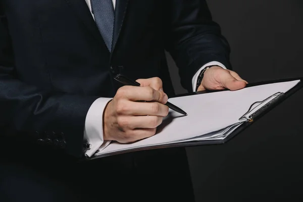 Vista cortada de empresário escrevendo em papel enquanto segurando prancheta isolada em preto — Fotografia de Stock