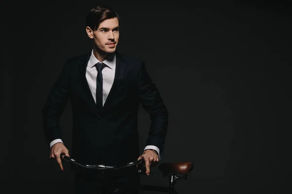 Guapo hombre de negocios en traje sosteniendo bicicleta aislado en negro - foto de stock