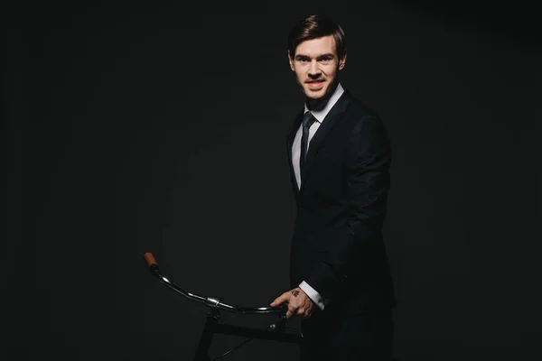 Smiling businessman in suit holding bicycle isolated on black — Stock Photo