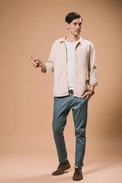 Bel homme tenant des lunettes debout avec la main dans la poche sur fond beige — Photo de stock