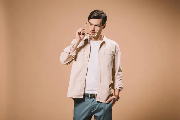 Hombre confiado con gafas y de pie con la mano en el bolsillo aislado sobre fondo beige - foto de stock