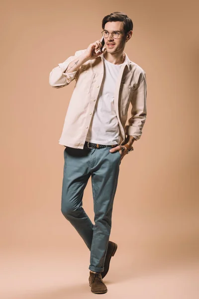 Happy man in glasses talking on smartphone while standing with hand in pocket on beige  background — Stock Photo