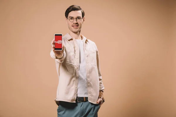 Allegro uomo in piedi con mano in tasca e tenendo smartphone con app youtube sullo schermo isolato su beige — Foto stock