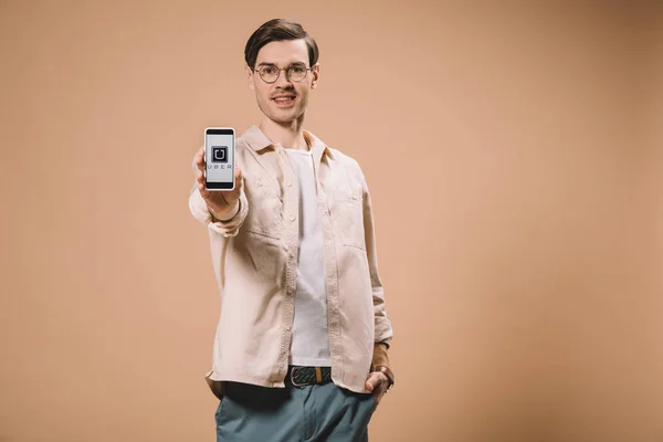 Fröhlicher Mann, der mit der Hand in der Tasche steht und Smartphone mit Uber-App isoliert auf dem Bildschirm in Beige hält — Stockfoto