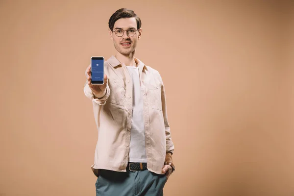 Homem alegre de pé com a mão no bolso e segurando smartphone com aplicativo facebook na tela isolada no bege — Fotografia de Stock