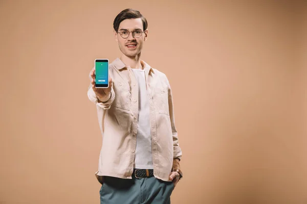 Hombre alegre de pie con la mano en el bolsillo y la celebración de teléfono inteligente con la aplicación de Twitter en la pantalla aislada en beige - foto de stock