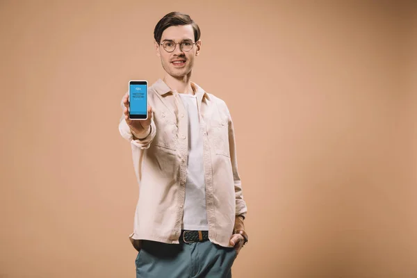 Hombre alegre de pie con la mano en el bolsillo y la celebración de teléfono inteligente con aplicación skype en solapa aislado en beige - foto de stock