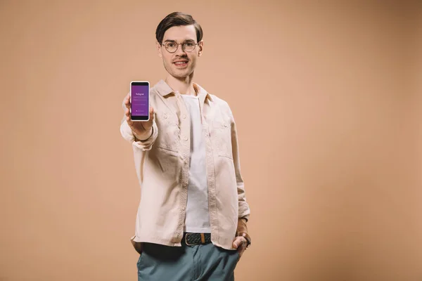Allegro uomo in piedi con mano in tasca e tenendo smartphone con app instagram sullo schermo isolato su beige — Foto stock