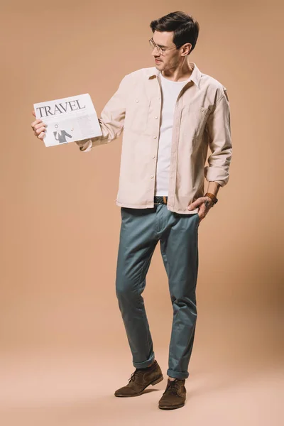 Hombre guapo con la mano en el bolsillo mirando el periódico de viaje sobre fondo beige - foto de stock