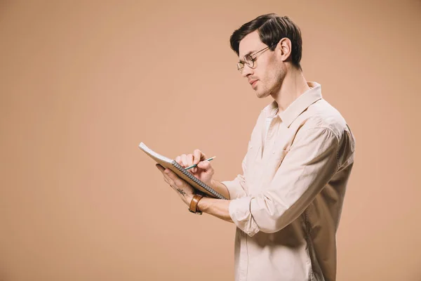 Thoughtful man in glasses writing notebook isolated on beige — Stock Photo